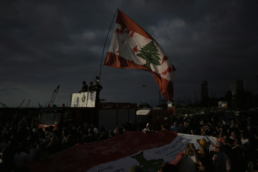 Familiares de las víctimas de la mortal explosión del puerto de Beirut en 2020 se encuentran en la escalera de un camión de bomberos, sosteniendo retratos de sus seres queridos y una bandera libanesa gigante, durante una reunión para conmemorar el quinto aniversario de la enorme explosión, a las afueras del puerto de Beirut, Líbano, el lunes 4 de agosto de 2025. (Foto AP)
