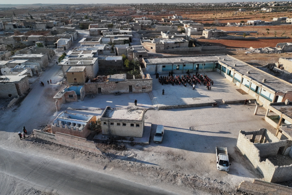 Estudiantes sirios se reúnen en el patio de la escuela primaria Maar Shmarin, rodeada de casas destruidas en Idlib, Siria